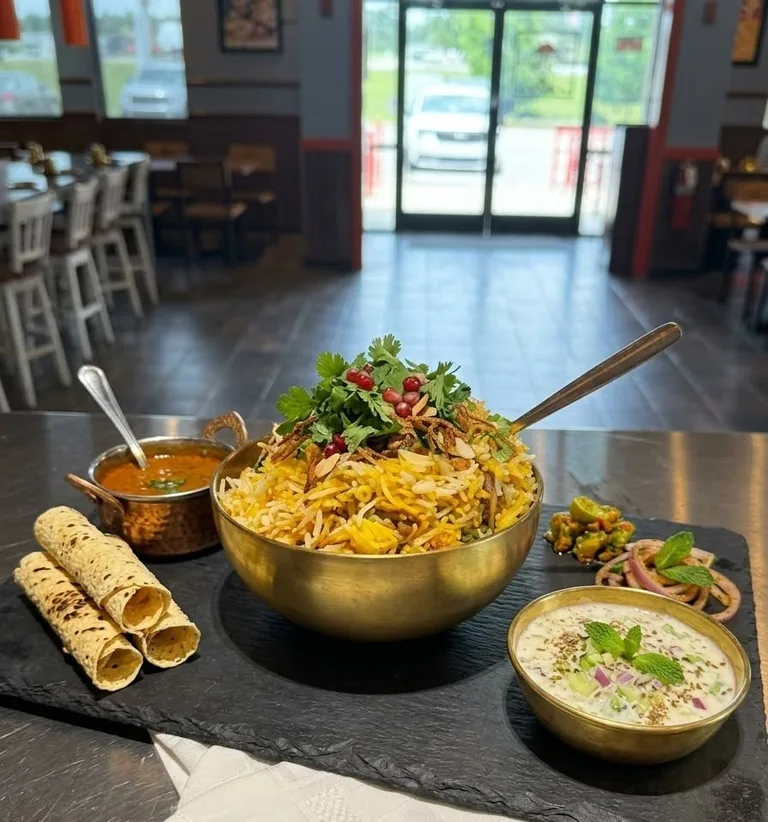 Lucknowi biryani in a gold bowl with a copper handi of curry, raita and papad rolls beside it.