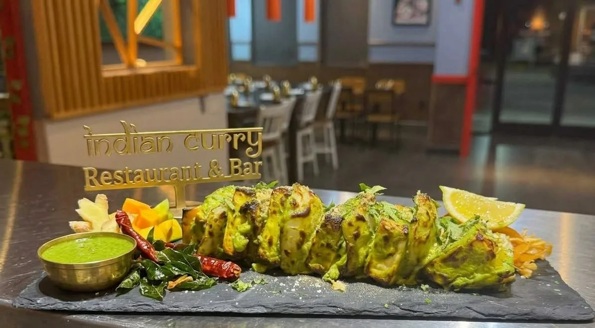 Green pesto paneer tikka on a black slate beside a gold sign reading indian curry Restaurant & Bar, the dining room behind.