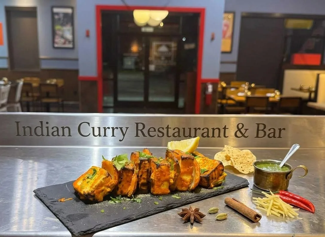 Paneer tikka on a black slate on the steel pass at Indian Curry, the restaurant's name engraved in the steel above it and the red front door beyond.