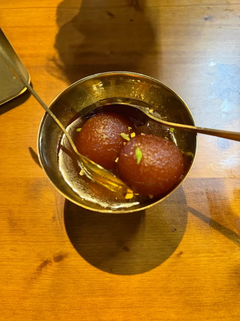 Two gulab jamun in a brass katori in their syrup, scattered with pistachio, gold spoons resting in the bowl.