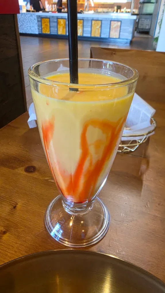 A mango lassi with a red syrup swirl, a gold plate edge in frame and the onyx counter behind.