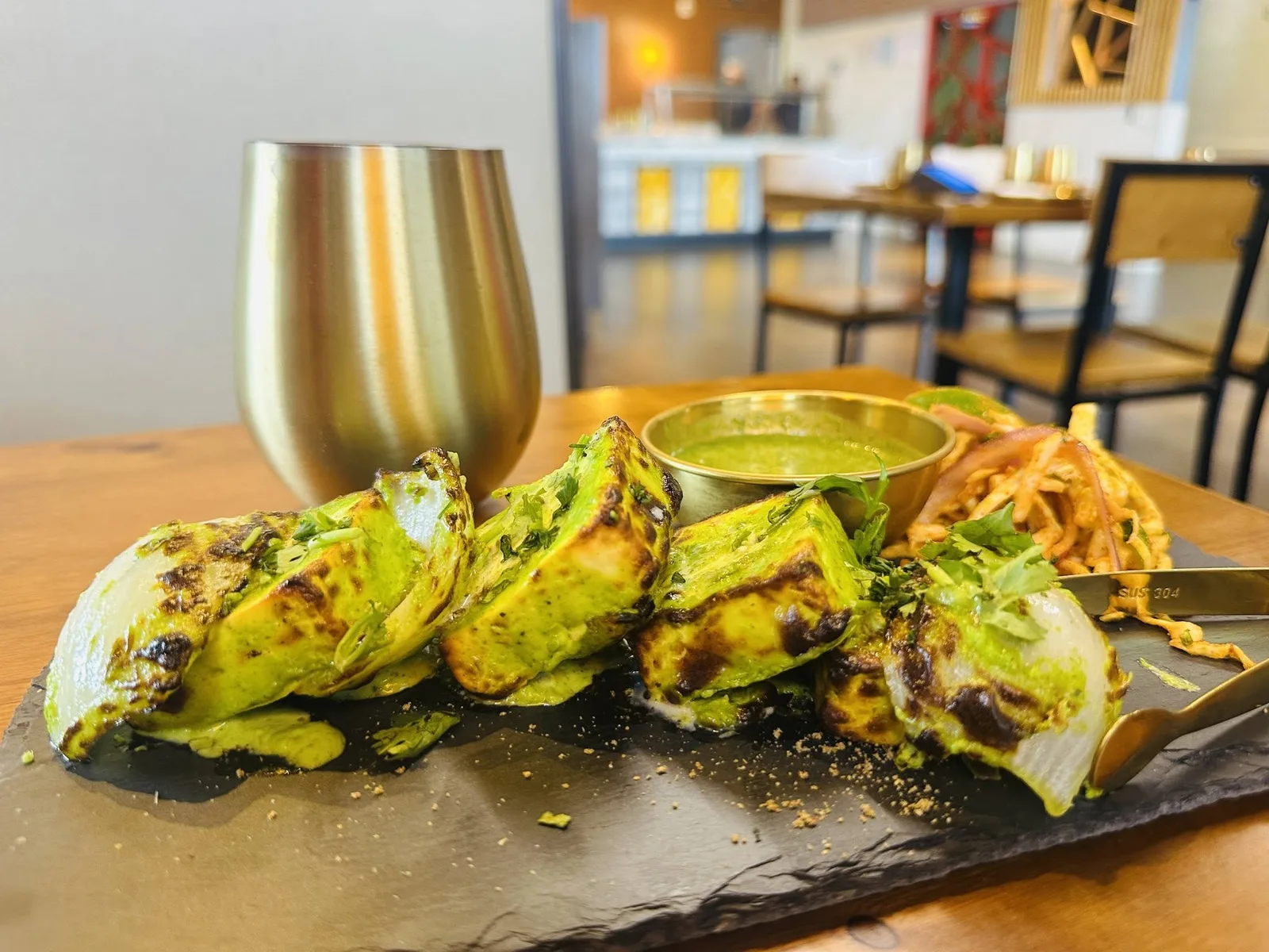 Hariyali paneer tikka, green with mint and coriander, on a black slate with a gold tumbler beside it.