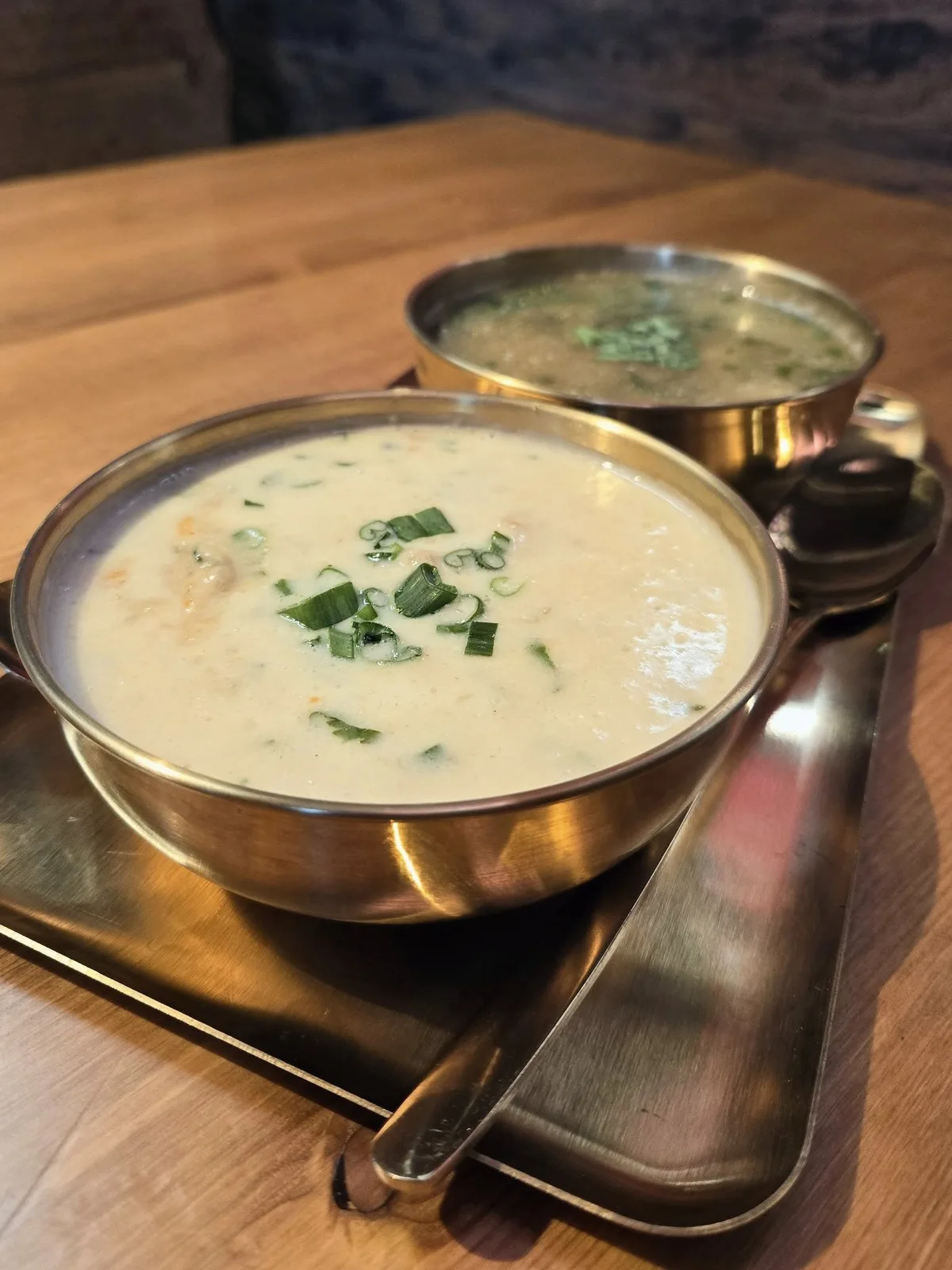 Sweet corn soup in a brass katori on a gold tray, a second katori and brass spoons behind.