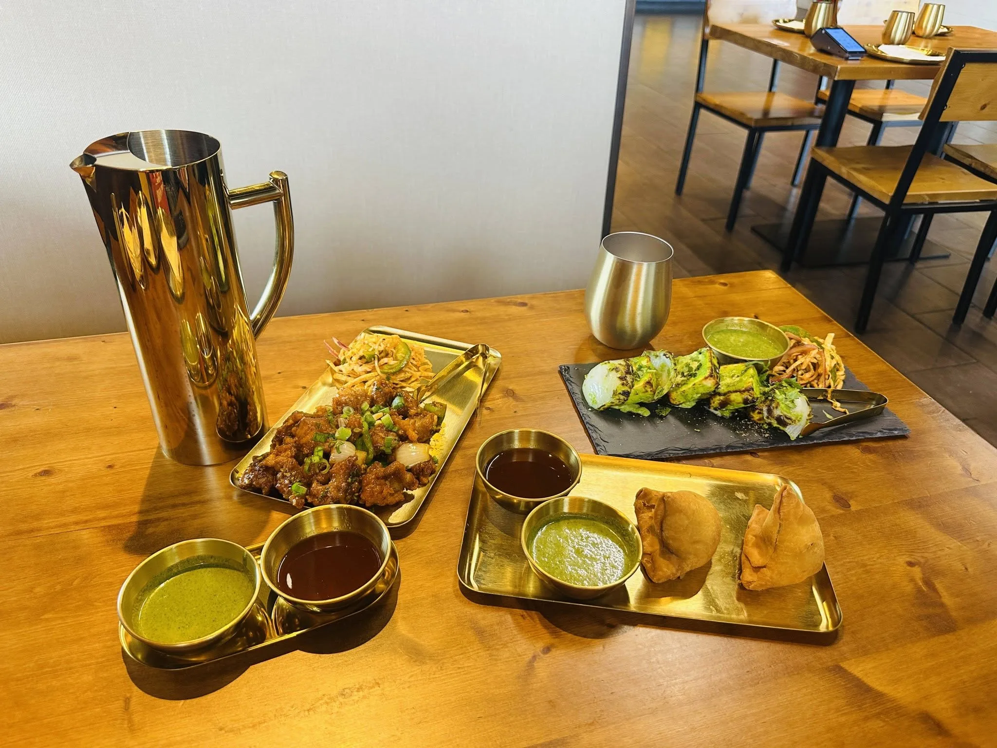The table at Indian Curry: a brass pitcher, chili chicken and hariyali tikka on slate, samosas and four brass katoris on a gold tray.