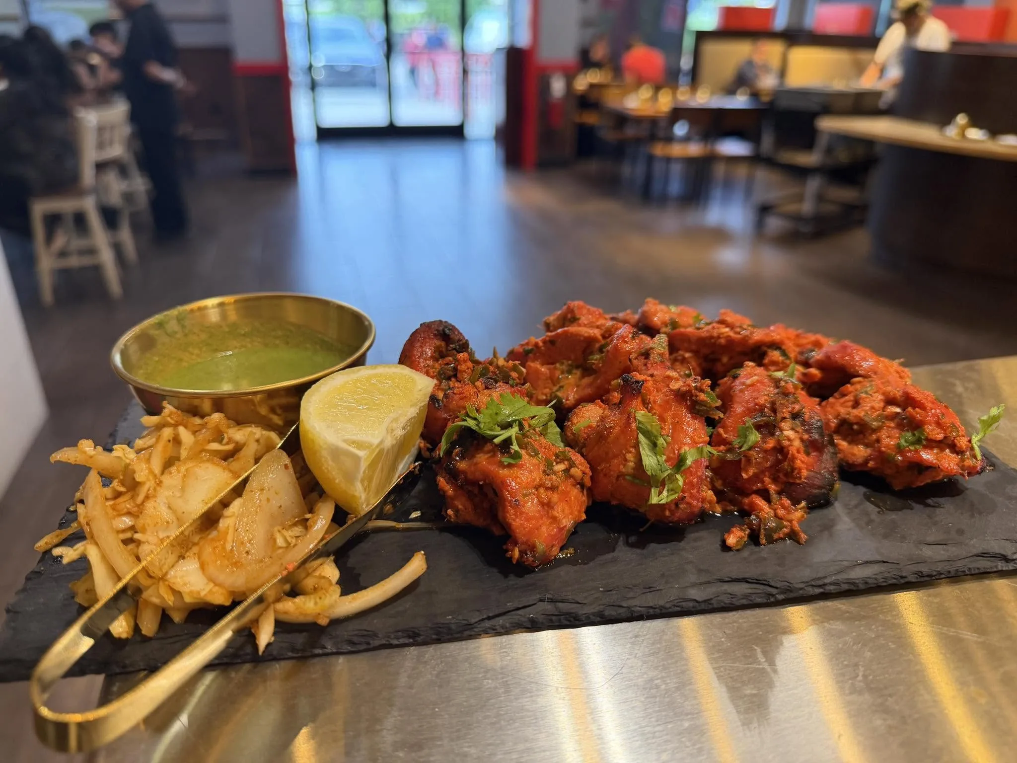 Tandoori chicken on a black slate with gold tongs, the dining room and front door behind.
